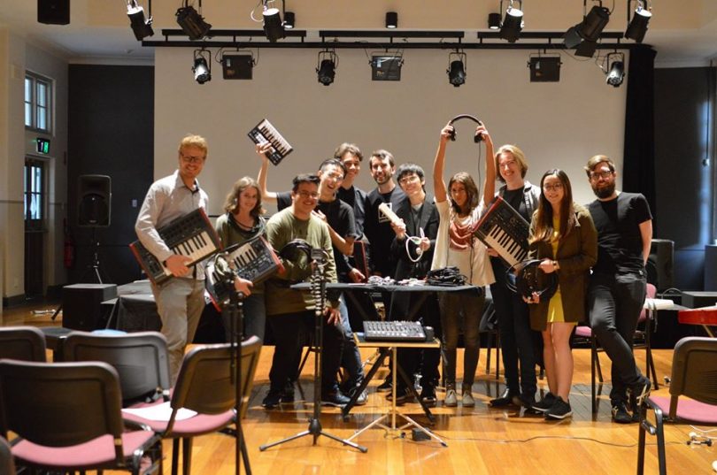 Photograph shows 10 members of ANU Laptop Ensemble celebrating with their laptops in a community hall